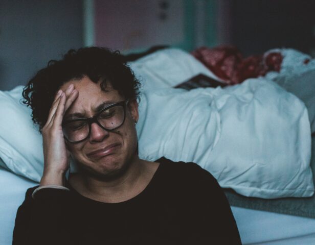 Woman sits on the floor beside the white bed, laying the heads of her right hand and crying