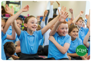 Primary school children in blue uniforms sitting on the floor and eagerly raising their hands to participate in a lesson