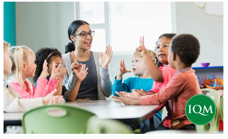 Teacher in classroom sitting at a desk full of children interacting with the teacher and each other