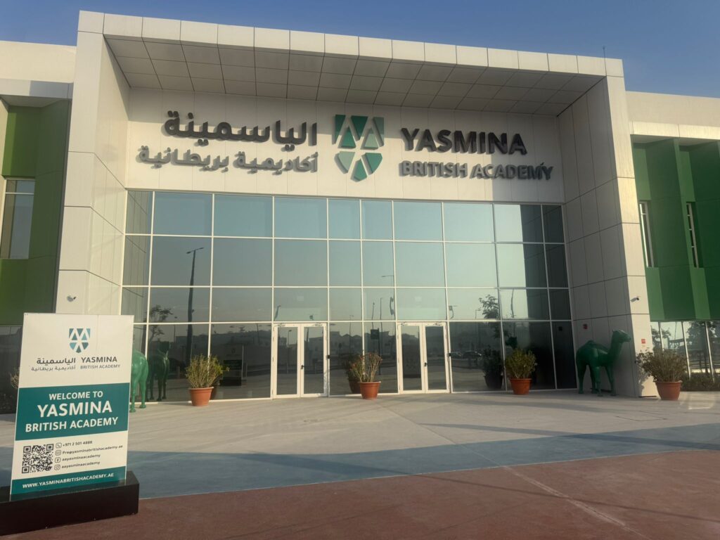 Yasmina British Academy school building, with large glass windows in the centre and plants in front. Two green camels either side of entrance.